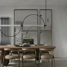 Load image into Gallery viewer, Solari Arc Light with brass arc and linear LED tube, sculptural pendant available in gold or black finish, pair of Solari Arc Lights hanging in modern dining space, minimalist arc pendant styled in office interior, contemporary pendant lighting with sleek LED tube, statement arc light fixture for high ceiling interiors. Modern dining room with wooden table and chairs, minimalistic decor, and abstract wall art.