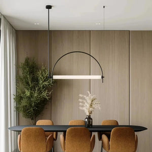Solari Arc Light with brass arc and linear LED tube, sculptural pendant available in gold or black finish, pair of Solari Arc Lights hanging in modern dining space, minimalist arc pendant styled in office interior, contemporary pendant lighting with sleek LED tube, statement arc light fixture for high ceiling interiors. Modern dining room with a black table, orange chairs, and a hanging light fixture.