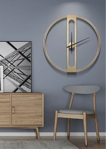 INSPIRA LIFESTYLES - Xavier Wall Clock, extra large wall clock, modern iron wall clock, sculptural wall decor, gold wall clock, charcoal wall clock, minimalist timepiece, statement wall accent, living room clock, office wall clock, oversized modern clock Modern wall clock on a gray wall with a wooden sideboard and chair in the foreground.