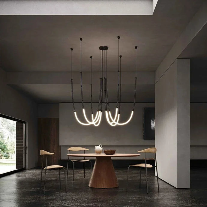 Siliq Sculptural Light with handcrafted silicone forms and metallic segmented cables, single minimalist pendant with glowing silicone tube, dramatic chandelier version of Siliq Light for high ceilings, floor-to-ceiling sculptural lighting installation in modern interior, flexible silicone light tubes emitting soft ambient glow, artistic designer light fixture styled in dining room, contemporary sculptural lighting as statement piece in commercial space.