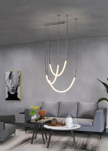 Load image into Gallery viewer, Siliq Sculptural Light with handcrafted silicone forms and metallic segmented cables, single minimalist pendant with glowing silicone tube, dramatic chandelier version of Siliq Light for high ceilings, floor-to-ceiling sculptural lighting installation in modern interior, flexible silicone light tubes emitting soft ambient glow, artistic designer light fixture styled in dining room, contemporary sculptural lighting as statement piece in commercial space.
