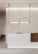 Load image into Gallery viewer, Solari Arc Light with brass arc and linear LED tube, sculptural pendant available in gold or black finish, pair of Solari Arc Lights hanging in modern dining space, minimalist arc pendant styled in office interior, contemporary pendant lighting with sleek LED tube, statement arc light fixture for high ceiling interiors. Modern interior design with a white reception desk and gold pendant light.
