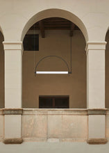 Load image into Gallery viewer, Solari Arc Light with brass arc and linear LED tube, sculptural pendant available in gold or black finish, pair of Solari Arc Lights hanging in modern dining space, minimalist arc pendant styled in office interior, contemporary pendant lighting with sleek LED tube, statement arc light fixture for high ceiling interiors. Architectural detail of a building with columns and an archway.
