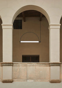 Solari Arc Light with brass arc and linear LED tube, sculptural pendant available in gold or black finish, pair of Solari Arc Lights hanging in modern dining space, minimalist arc pendant styled in office interior, contemporary pendant lighting with sleek LED tube, statement arc light fixture for high ceiling interiors. Architectural detail of a building with columns and an archway.