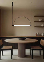 Load image into Gallery viewer, Solari Arc Light with brass arc and linear LED tube, sculptural pendant available in gold or black finish, pair of Solari Arc Lights hanging in modern dining space, minimalist arc pendant styled in office interior, contemporary pendant lighting with sleek LED tube, statement arc light fixture for high ceiling interiors. Modern dining room with a round wooden table and chairs, featuring a hanging light fixture.
