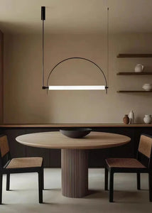 Solari Arc Light with brass arc and linear LED tube, sculptural pendant available in gold or black finish, pair of Solari Arc Lights hanging in modern dining space, minimalist arc pendant styled in office interior, contemporary pendant lighting with sleek LED tube, statement arc light fixture for high ceiling interiors. Modern dining room with a round wooden table and chairs, featuring a hanging light fixture.