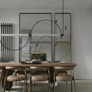 Solari Arc Light with brass arc and linear LED tube, sculptural pendant available in gold or black finish, pair of Solari Arc Lights hanging in modern dining space, minimalist arc pendant styled in office interior, contemporary pendant lighting with sleek LED tube, statement arc light fixture for high ceiling interiors. Modern dining room with wooden table and chairs, minimalistic decor, and abstract wall art.