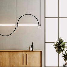 Load image into Gallery viewer, Solari Arc Light with brass arc and linear LED tube, sculptural pendant available in gold or black finish, pair of Solari Arc Lights hanging in modern dining space, minimalist arc pendant styled in office interior, contemporary pendant lighting with sleek LED tube, statement arc light fixture for high ceiling interiors. Modern pendant light fixture in a minimalistic room with wooden cabinet and plants.
