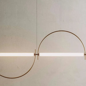 Solari Arc Light with brass arc and linear LED tube, sculptural pendant available in gold or black finish, pair of Solari Arc Lights hanging in modern dining space, minimalist arc pendant styled in office interior, contemporary pendant lighting with sleek LED tube, statement arc light fixture for high ceiling interiors. Modern wall light fixture with a curved design on a plain wall.