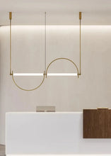 Load image into Gallery viewer, Solari Arc Light with brass arc and linear LED tube, sculptural pendant available in gold or black finish, pair of Solari Arc Lights hanging in modern dining space, minimalist arc pendant styled in office interior, contemporary pendant lighting with sleek LED tube, statement arc light fixture for high ceiling interiors. Modern pendant light fixture hanging above a reception desk in an office setting.
