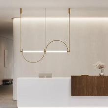 Load image into Gallery viewer, Solari Arc Light with brass arc and linear LED tube, sculptural pendant available in gold or black finish, pair of Solari Arc Lights hanging in modern dining space, minimalist arc pendant styled in office interior, contemporary pendant lighting with sleek LED tube, statement arc light fixture for high ceiling interiors. Modern interior design with a pendant light fixture and reception desk.
