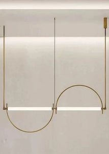 Solari Arc Light with brass arc and linear LED tube, sculptural pendant available in gold or black finish, pair of Solari Arc Lights hanging in modern dining space, minimalist arc pendant styled in office interior, contemporary pendant lighting with sleek LED tube, statement arc light fixture for high ceiling interiors. Modern pendant light fixture with curved design on a neutral background