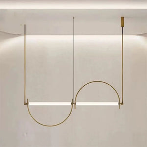 Solari Arc Light with brass arc and linear LED tube, sculptural pendant available in gold or black finish, pair of Solari Arc Lights hanging in modern dining space, minimalist arc pendant styled in office interior, contemporary pendant lighting with sleek LED tube, statement arc light fixture for high ceiling interiors. Modern pendant light fixture with a curved design on a neutral background