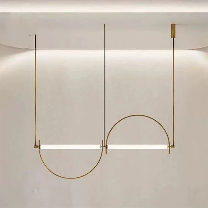 Solari Arc Light with brass arc and linear LED tube, sculptural pendant available in gold or black finish, pair of Solari Arc Lights hanging in modern dining space, minimalist arc pendant styled in office interior, contemporary pendant lighting with sleek LED tube, statement arc light fixture for high ceiling interiors. Modern pendant light fixture with a curved design on a neutral background