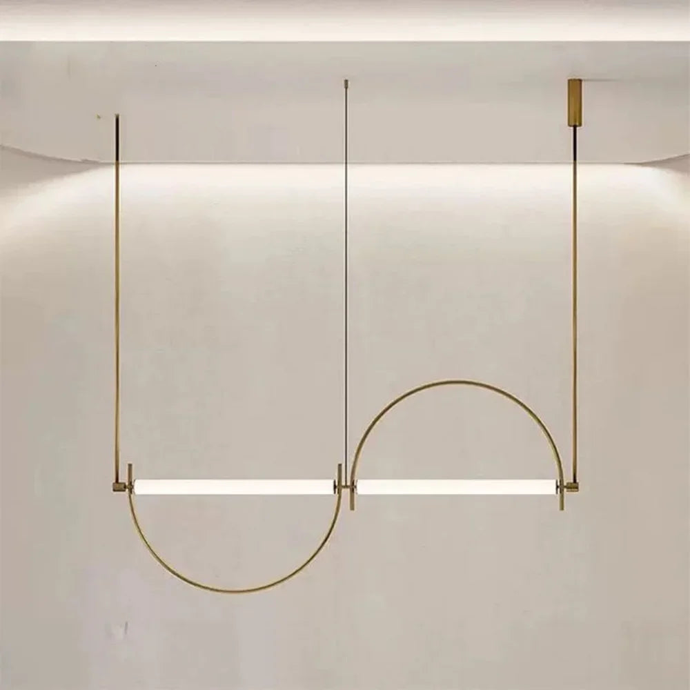 Solari Arc Light with brass arc and linear LED tube, sculptural pendant available in gold or black finish, pair of Solari Arc Lights hanging in modern dining space, minimalist arc pendant styled in office interior, contemporary pendant lighting with sleek LED tube, statement arc light fixture for high ceiling interiors. Modern pendant light fixture with a curved design on a neutral background