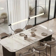Load image into Gallery viewer, Solari Arc Light with brass arc and linear LED tube, sculptural pendant available in gold or black finish, pair of Solari Arc Lights hanging in modern dining space, minimalist arc pendant styled in office interior, contemporary pendant lighting with sleek LED tube, statement arc light fixture for high ceiling interiors. Modern dining table with marble surface and chairs, featuring a stylish pendant light.
