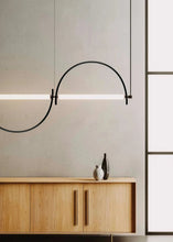 Load image into Gallery viewer, Solari Arc Light with brass arc and linear LED tube, sculptural pendant available in gold or black finish, pair of Solari Arc Lights hanging in modern dining space, minimalist arc pendant styled in office interior, contemporary pendant lighting with sleek LED tube, statement arc light fixture for high ceiling interiors. Modern pendant light fixture hanging above a wooden cabinet with a neutral wall background.
