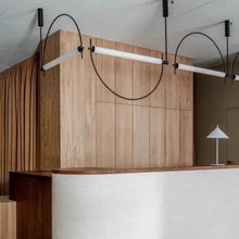 Load image into Gallery viewer, Solari Arc Light with brass arc and linear LED tube, sculptural pendant available in gold or black finish, pair of Solari Arc Lights hanging in modern dining space, minimalist arc pendant styled in office interior, contemporary pendant lighting with sleek LED tube, statement arc light fixture for high ceiling interiors. Modern interior with wooden paneling and minimalist lighting fixtures.
