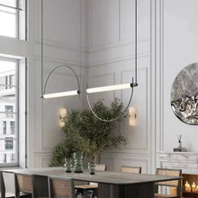 Load image into Gallery viewer, Solari Arc Light with brass arc and linear LED tube, sculptural pendant available in gold or black finish, pair of Solari Arc Lights hanging in modern dining space, minimalist arc pendant styled in office interior, contemporary pendant lighting with sleek LED tube, statement arc light fixture for high ceiling interiors. Modern dining room with a long pendant light fixture, table, chairs, and decorative elements.
