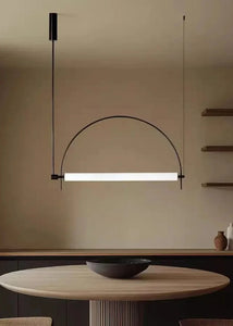 Solari Arc Light with brass arc and linear LED tube, sculptural pendant available in gold or black finish, pair of Solari Arc Lights hanging in modern dining space, minimalist arc pendant styled in office interior, contemporary pendant lighting with sleek LED tube, statement arc light fixture for high ceiling interiors. Modern pendant light hanging above a dining table with a neutral background