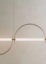 Load image into Gallery viewer, Solari Arc Light with brass arc and linear LED tube, sculptural pendant available in gold or black finish, pair of Solari Arc Lights hanging in modern dining space, minimalist arc pendant styled in office interior, contemporary pendant lighting with sleek LED tube, statement arc light fixture for high ceiling interiors. Modern wall-mounted light fixture with a curved design on a neutral background

