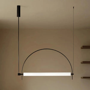 Solari Arc Light with brass arc and linear LED tube, sculptural pendant available in gold or black finish, pair of Solari Arc Lights hanging in modern dining space, minimalist arc pendant styled in office interior, contemporary pendant lighting with sleek LED tube, statement arc light fixture for high ceiling interiors. Modern pendant light with a curved design hanging against a neutral wall.