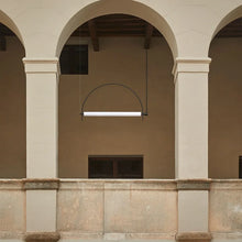 Load image into Gallery viewer, Solari Arc Light with brass arc and linear LED tube, sculptural pendant available in gold or black finish, pair of Solari Arc Lights hanging in modern dining space, minimalist arc pendant styled in office interior, contemporary pendant lighting with sleek LED tube, statement arc light fixture for high ceiling interiors. Architectural detail of a building with arches and a modern light fixture.
