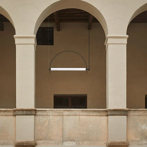 Solari Arc Light with brass arc and linear LED tube, sculptural pendant available in gold or black finish, pair of Solari Arc Lights hanging in modern dining space, minimalist arc pendant styled in office interior, contemporary pendant lighting with sleek LED tube, statement arc light fixture for high ceiling interiors. Architectural detail of a building with arches and a modern light fixture.