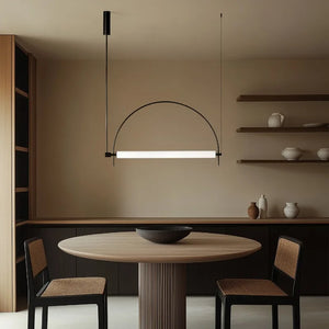 Solari Arc Light with brass arc and linear LED tube, sculptural pendant available in gold or black finish, pair of Solari Arc Lights hanging in modern dining space, minimalist arc pendant styled in office interior, contemporary pendant lighting with sleek LED tube, statement arc light fixture for high ceiling interiors. Modern dining area with a round table, chairs, and minimalist lighting fixture.