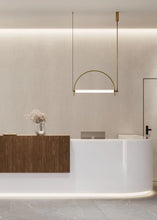 Load image into Gallery viewer, Solari Arc Light with brass arc and linear LED tube, sculptural pendant available in gold or black finish, pair of Solari Arc Lights hanging in modern dining space, minimalist arc pendant styled in office interior, contemporary pendant lighting with sleek LED tube, statement arc light fixture for high ceiling interiors. Modern interior design with a reception desk and pendant light fixture.
