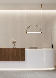 Solari Arc Light with brass arc and linear LED tube, sculptural pendant available in gold or black finish, pair of Solari Arc Lights hanging in modern dining space, minimalist arc pendant styled in office interior, contemporary pendant lighting with sleek LED tube, statement arc light fixture for high ceiling interiors. Modern interior design with a reception desk and pendant light fixture.