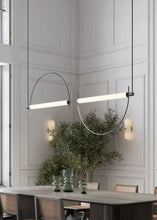 Load image into Gallery viewer, Solari Arc Light with brass arc and linear LED tube, sculptural pendant available in gold or black finish, pair of Solari Arc Lights hanging in modern dining space, minimalist arc pendant styled in office interior, contemporary pendant lighting with sleek LED tube, statement arc light fixture for high ceiling interiors. Modern dining room with a long pendant light fixture, table, chairs, and decorative elements.
