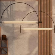 Load image into Gallery viewer, Solari Arc Light with brass arc and linear LED tube, sculptural pendant available in gold or black finish, pair of Solari Arc Lights hanging in modern dining space, minimalist arc pendant styled in office interior, contemporary pendant lighting with sleek LED tube, statement arc light fixture for high ceiling interiors. Modern pendant light fixture with curved metal arm and cylindrical bulbs against a textured wall.
